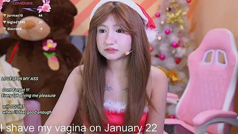 Media: Video of an Asian woman with long brown hair, wearing a red Santa hat, and a strapless red dress, in a Christmas-themed room, with a pink gaming chair. Text overlay reads \"I share my vagina on January 22.\