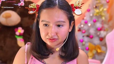 Media: Video of a young Asian girl with straight black hair styled with gold and red bows, wearing a pink dress, against a festive background with a Christmas tree and decorations.