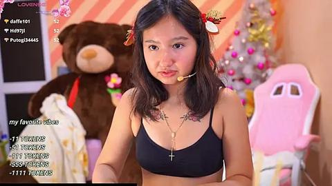 Media: A video of an East Asian woman with shoulder-length black hair in a black bra, wearing a microphone headset, seated in a pink gaming chair. Background includes a stuffed bear and a decorated Christmas tree.