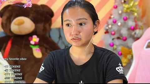 Media: Video of an Asian woman with medium skin tone and dark hair, wearing a black t-shirt, looking serious. Background features a large stuffed bear and festive decorations.