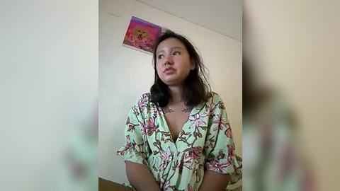 Media: A video of a young East Asian woman with medium-length black hair, wearing a green floral robe, standing in a plain, cream-colored room with a colorful abstract painting on the wall.