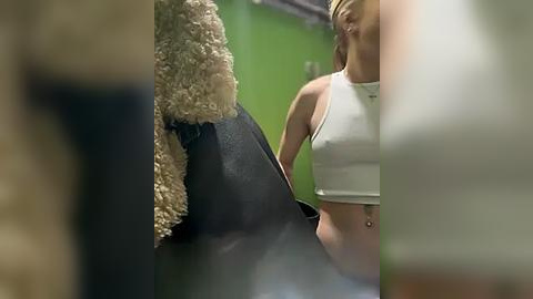 Media: Video of a blonde woman in a white crop top and shorts, holding a fluffy, beige cat, in a dimly lit room with green walls.