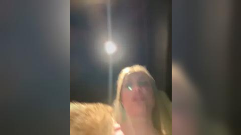 Media: A blurry video of a blonde woman with a fur coat, wearing glasses, looking up at a bright light source. The background is dark and indistinct.