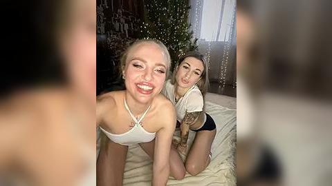 Media: Video of two young, smiling, blonde women in white crop tops, posing playfully on a bed with a beige blanket.