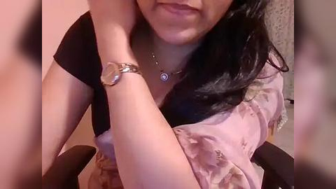 Media: Video of a woman with medium skin tone, long black hair, wearing a light pink floral blouse, black arm, and silver watch. Background is blurred.