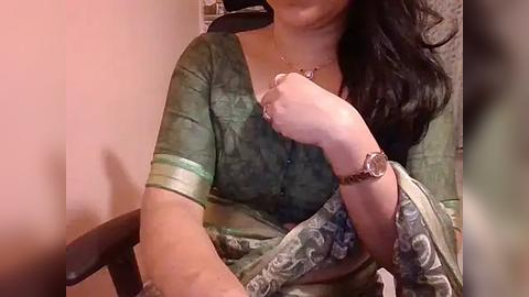 Media: Video of an Indian woman with medium skin tone and long black hair, wearing a green blouse with a floral pattern and a white sari, seated in a chair, holding her wrist, with a blurred background.