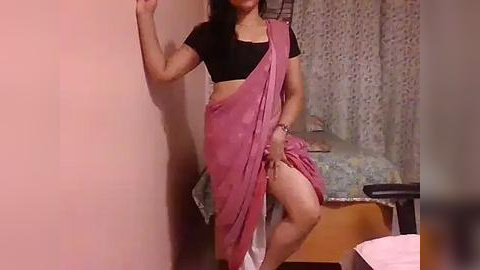 Media: A video of a South Asian woman in a black crop top and pink saree, striking a pose in a modest, dimly-lit room with floral curtains and a bed.