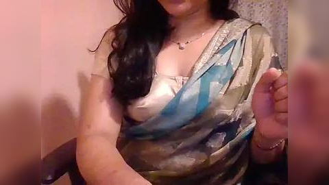 Media: Video of a woman with long dark hair, wearing a blue and beige sari, raising her right hand in a casual pose. She has a light skin tone, and the background is blurred.