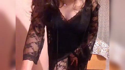 Media: Video of a woman in black lace lingerie with sheer sleeves, standing indoors, her long, wavy hair cascading over her shoulders.