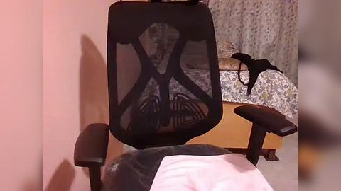 Media: A video of a modern black office chair with a mesh backrest, positioned in a cozy bedroom with a patterned curtain and a bed with a floral duvet.