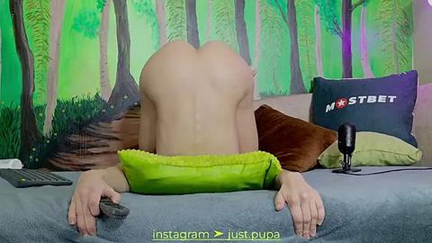 Media: Video of a nude woman with fair skin, facing away, lying on a bed with green pillows and a brown cushion, in a room with a forest mural.