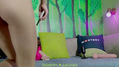 Media: Video of a nude woman with light skin, holding a dildo, standing beside a bed with a green pillow and a \"Mistress\" pillow, in a room with a green forest mural and purple lighting.