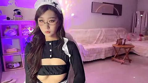 Media: A video of an Asian woman in a revealing black maid outfit with a white apron and headband, posing in a cozy, dimly lit living room with a TV, shelves, and plush toys.