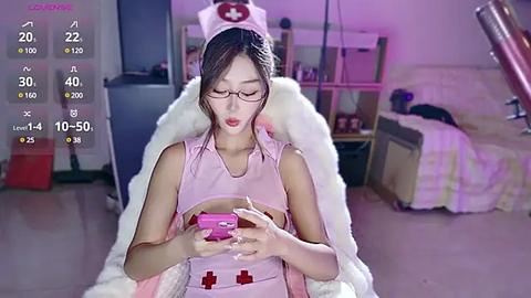 Media: Video of an East Asian woman in a pink nurse outfit with a red cross cap, sitting on a white chair, holding a pink box, in a dimly lit room with a bed and shelves in the background.