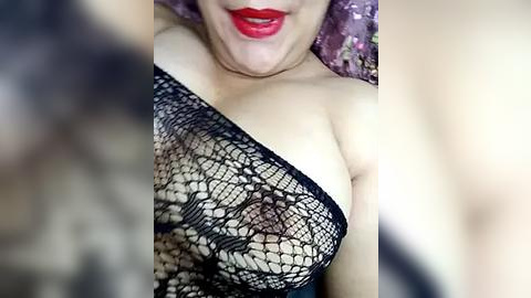 Media: Video of a woman with fair skin, wearing a black fishnet bodysuit that reveals her breasts, with red lipstick and a floral patterned bedsheet in the background.