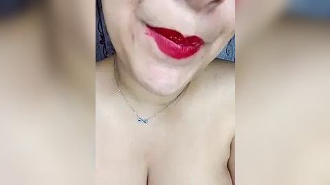 Media: Close-up video of a fair-skinned woman with full red lips, wearing a silver necklace with a small pendant, and a hint of her bare shoulders. Background is blurred.