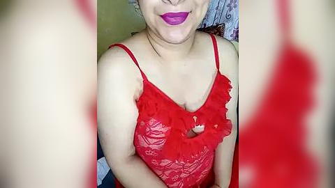 Media: Video of a light-skinned woman with a full smile, wearing a bright red lace camisole, with a visible heart cutout at the chest. Background features a blurred, patterned wall.