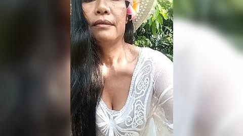 Media: Video of a smiling woman with long black hair and a light complexion, wearing a white lace top, standing outdoors in a lush green garden.