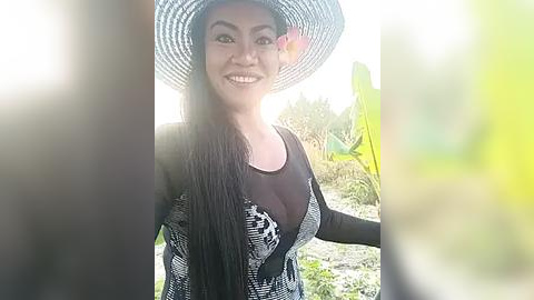Media: Video of a smiling Southeast Asian woman with long black hair, wearing a wide-brimmed straw hat adorned with a pink flower, black top, and checkered vest, standing outdoors among cacti and green foliage.