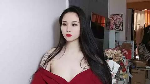 Media: Video of an East Asian woman with long black hair, red lipstick, and a red dress, standing in a cluttered room with a TV, flowers, and boxes.