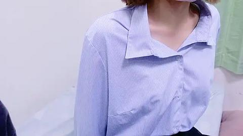 Media: Video of a light-skinned woman with short brown hair, wearing a light blue, button-down shirt with horizontal stripes, sitting indoors against a plain white wall.