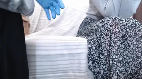 Media: Video of a person in a white lab coat and blue gloves adjusting a white, striped mattress cover on a bed with a black, floral-patterned bedspread.