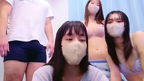 Media: Video of three Asian women wearing surgical masks, one in a white top, another in a pink bra, and the third in a white sweater, in a clinical setting with blue curtains.