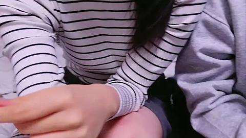 Media: Video of a person with pale skin and long black hair wearing a white and black striped long-sleeve shirt, cradling a baby's bottom, with a gray blanket in the background.