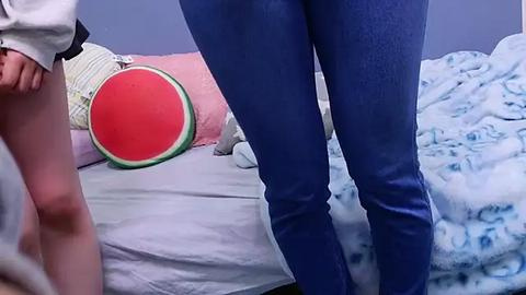 Media: Video of a person in blue leggings, standing by a bed with a watermelon pillow and a blue-patterned blanket.