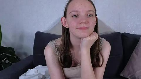 Media: Video of a young Caucasian woman with long brown hair, fair skin, and a slight smile, resting her chin on her hand on a dark blue couch, with a green plant and white pillow in the background.