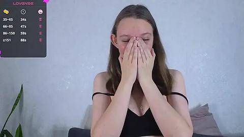 Media: Video of a fair-skinned, brunette woman with long hair, covering her face with her hands, wearing a black spaghetti-strap top. Background includes a gray wall, a potted plant, and a bed with gray pillows.