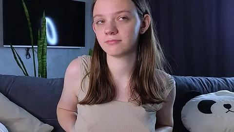 Media: Video of a young Caucasian girl with straight, shoulder-length brown hair, wearing a beige tank top, sitting on a gray couch. Background features a green plant, a panda plush toy, and a dark curtain.