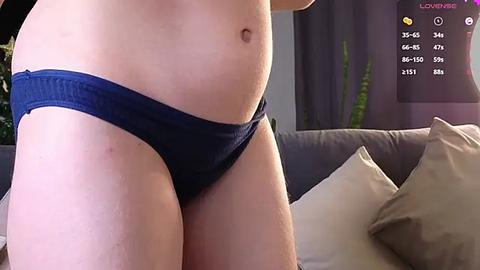 Media: Video of a fair-skinned person's lower torso in navy blue underwear, lying on a beige couch with pillows. Background includes a green plant and a TV screen displaying social media stats.