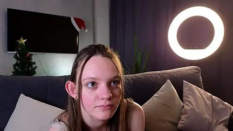 Media: A video of a young white woman with long brown hair, wearing a sleeveless top, sitting on a gray couch with pillows. A round lightbulb and a Christmas tree are visible in the background.