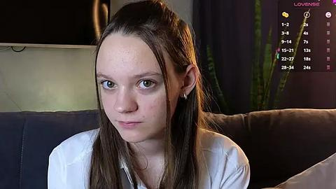 Media: A video of a young woman with fair skin and straight brown hair, sitting on a couch, wearing a white shirt. Background shows a dark room with a flat-screen TV and green plants.