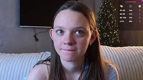 Media: Video of a young Caucasian woman with light skin, straight brown hair, and blue eyes, wearing a gray shirt, sitting on a white couch in a cozy living room with a TV and a Christmas tree.