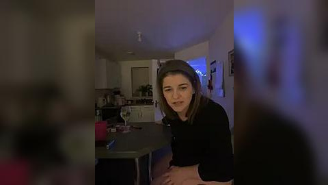 Media: Video of a woman with straight, shoulder-length brown hair, wearing a black top, sitting at a kitchen table. Background shows a dimly lit kitchen with white cabinets, a counter, and a blue light in the corner.