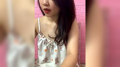 Media: Video of an East Asian woman with long black hair and light skin, wearing a white tank top with cartoon bear patterns, seated against a pink brick wall.