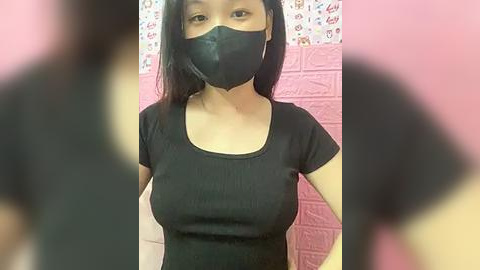 Media: Video of an Asian woman with straight black hair, wearing a black face mask, black t-shirt, and pink brick wall background.