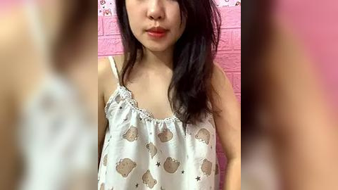 Media: Video of a young Asian woman with long black hair, wearing a white tank top with bear pattern, against a pink background.