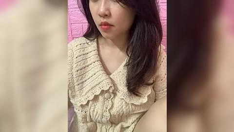 Media: Video of an East Asian woman with long black hair, fair skin, and red lipstick, wearing a cream-colored crocheted top with ruffled edges. Background features a pink brick wall.