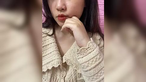 Media: Video of an Asian woman with long dark hair, wearing a beige, crocheted cardigan, resting her chin on her hand, against a pink background.