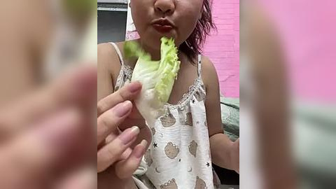 Media: Video of a young Asian girl with medium skin tone, wearing a white polka-dot dress, eating a green leafy vegetable. The background shows a pink wall and a blurred person.