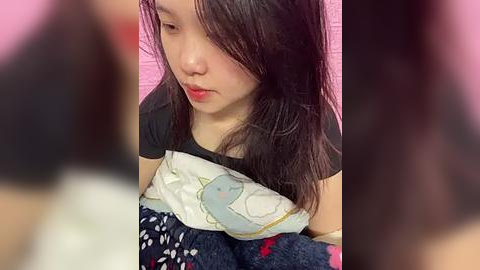 Media: A video of a young East Asian girl with long black hair, wearing a black t-shirt and blue pajama pants, lying on a white pillow with cartoon characters. Background is a blurred pink wall.