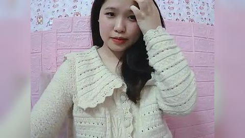 Media: A video of an Asian woman with straight black hair, wearing a beige crochet cardigan, standing against a pink wall with a patterned border. She is adjusting her hair with her right hand.