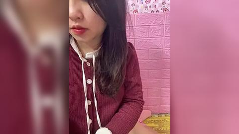 Media: Video of a young woman with long black hair and fair skin, wearing a red cardigan and white blouse, sitting on a pink bed with floral bedding, in a pink room with textured walls.