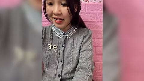 Media: Video of an Asian girl with straight dark hair, wearing a gray checkered dress with a pearl necklace, standing in front of a pink brick wall.