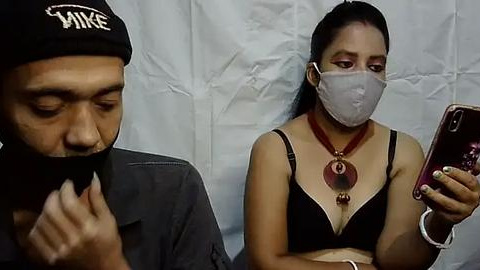 Media: Video of a man with a beard and black beanie, and a woman with a mask, necklace, and black bra, both taking selfies.