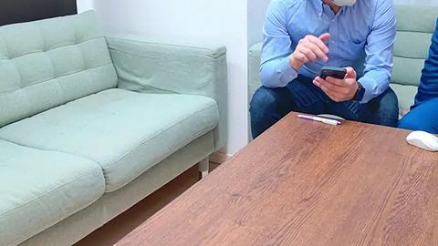 Media: Video of a man in a blue button-up shirt and jeans, sitting on a light green couch, holding a smartphone, in a modern living room with a wooden coffee table and white walls.