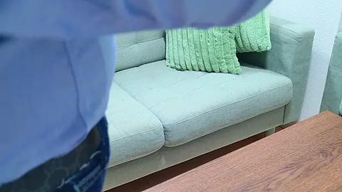 Media: Video of a light green, textured sofa with two green, ribbed pillows, viewed from a low angle. Person in blue shirt partially visible in foreground, standing on wooden floor.